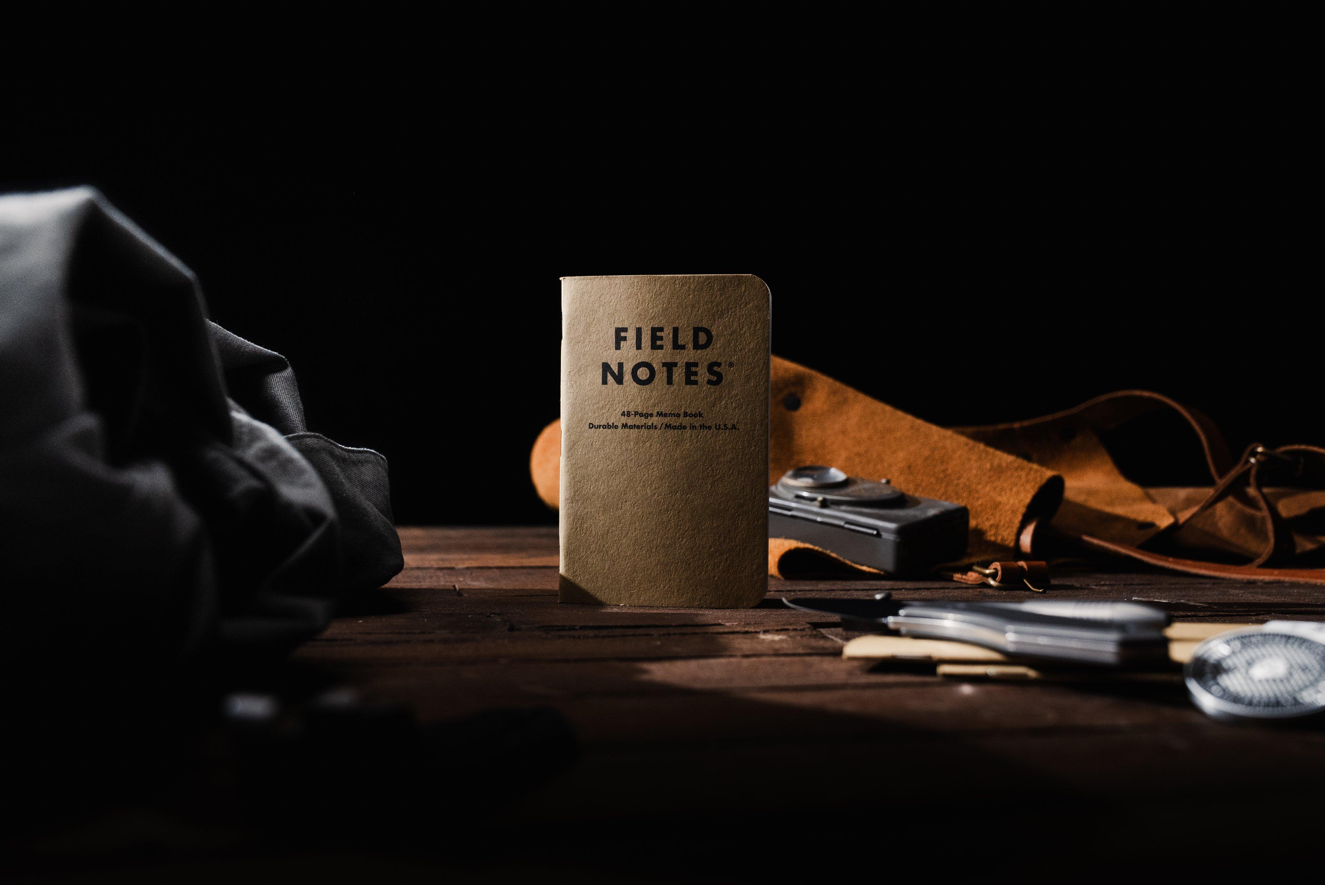 Un cuadero Field Notes sobre una mesa de madera, rodeado de un maletín de cuero, una cámara y otros accesorios de viaje en un entorno iluminado ligeramente.