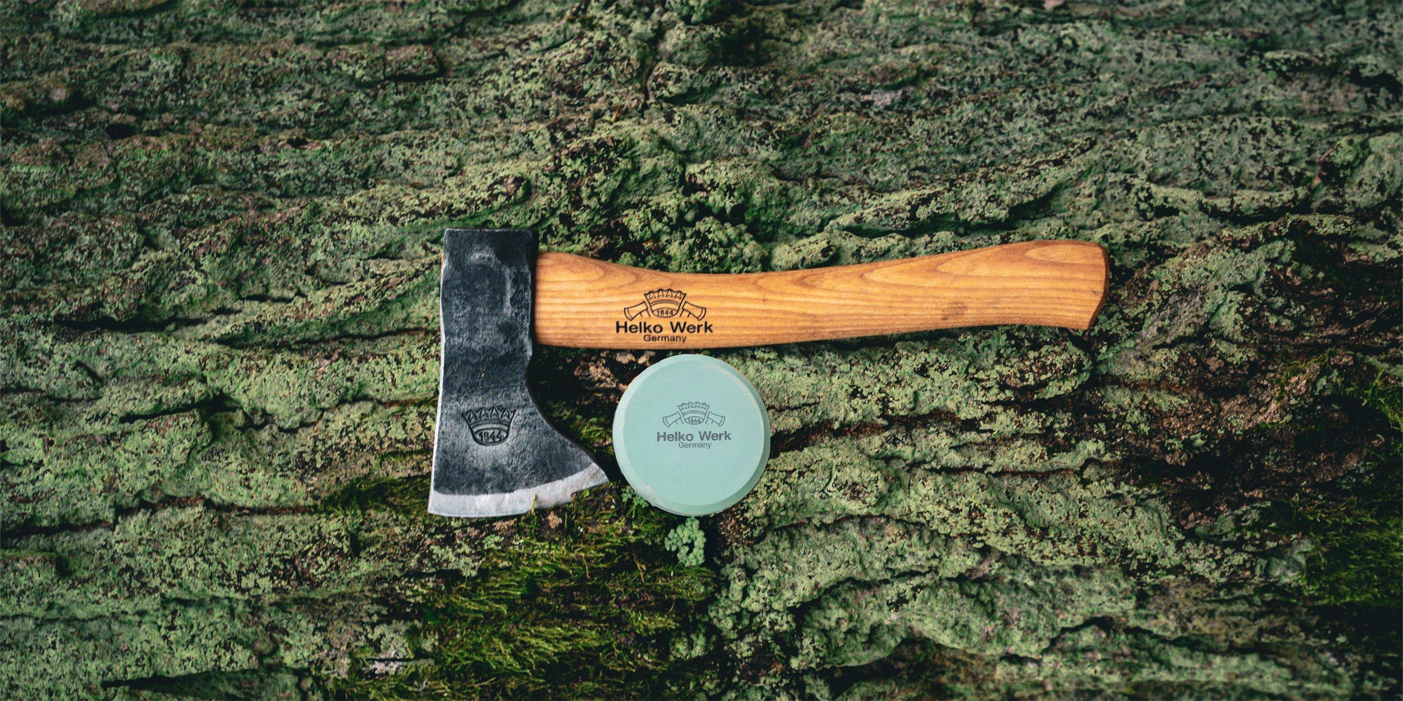 Un hacha con mango de madera y el logo de Helko Werk descansa sobre un tronco cubierto de musgo, junto a una piedra de afilar redonda.
