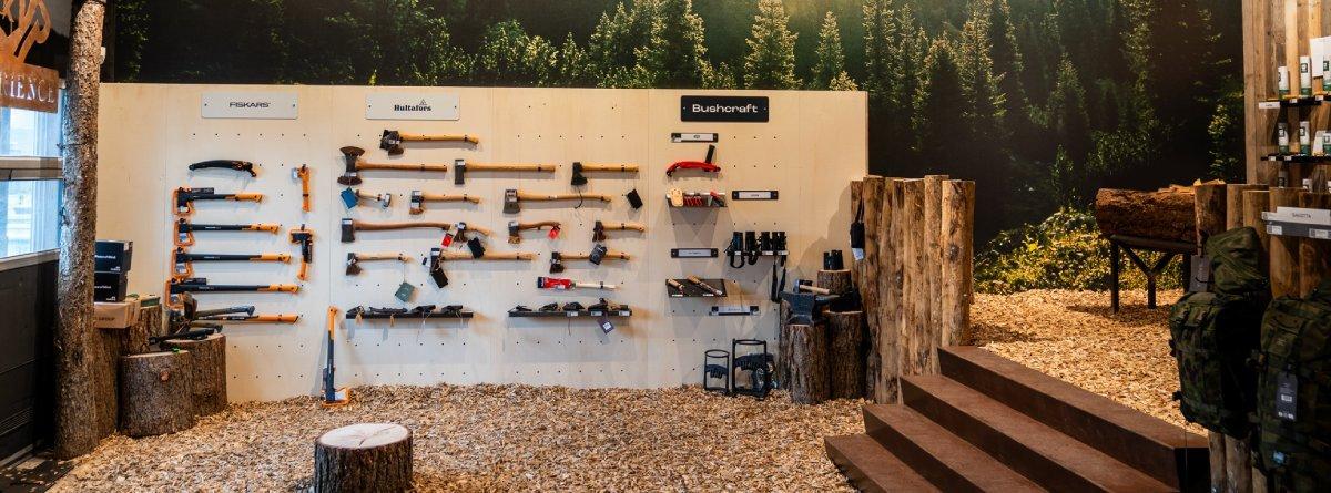 Äste und Beile verschiedener Hersteller auf Regalen im Knivesandtools Experience Center umrahmt von Holzstämmen und einer Tapete, die einen Wald zeig