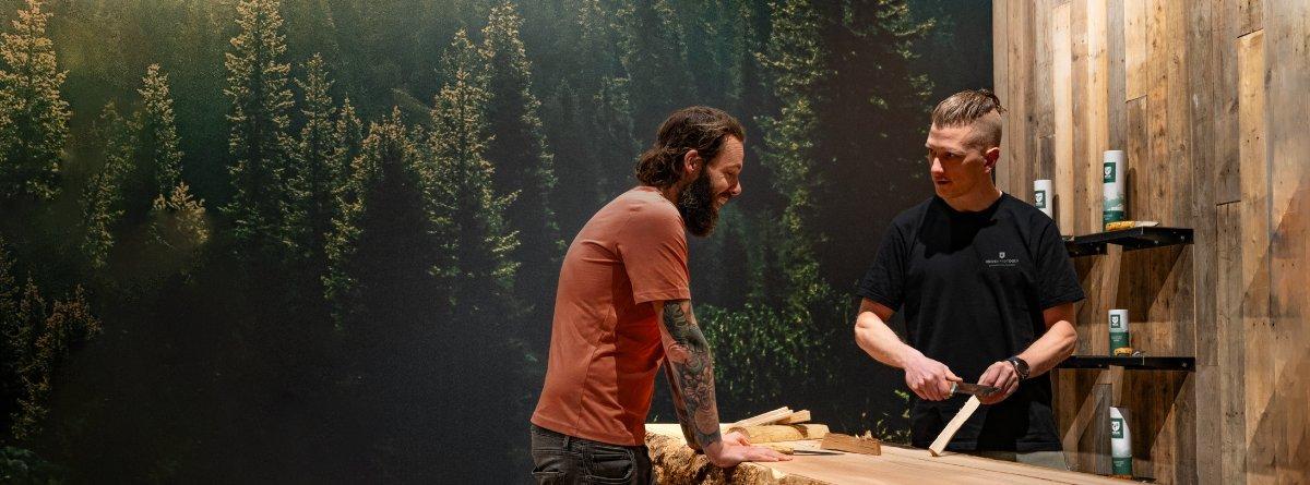 Ein Mann mit orangefarbenem Shirt langen dunklen Haaren, Bart und tätowierten Armen wird von einem Mitarbeiter von Knivesandtools im Knivesandtools Experience Center beraten. Ihm wird gezeigt, wie man mit einem Fahrtenmesser Feathersticks herstellt.