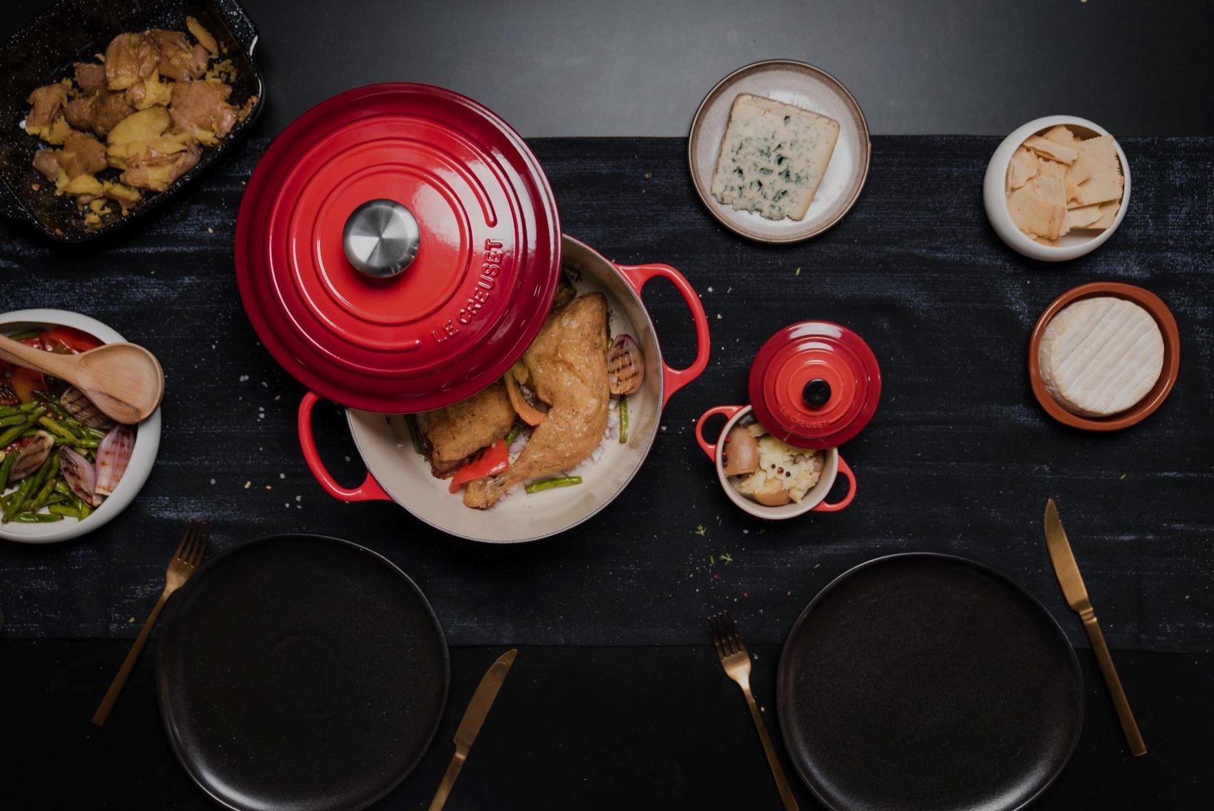 Ein roter Le Creuset Bräter und eine Mini-Cocotte auf einer dunklen Tischdecke.