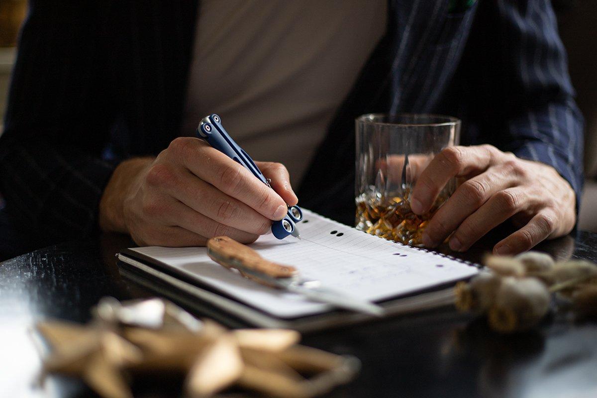 Alguien escribiendo en un cuaderno con un bolígrafo mientras disfruta de un vaso de whisky.