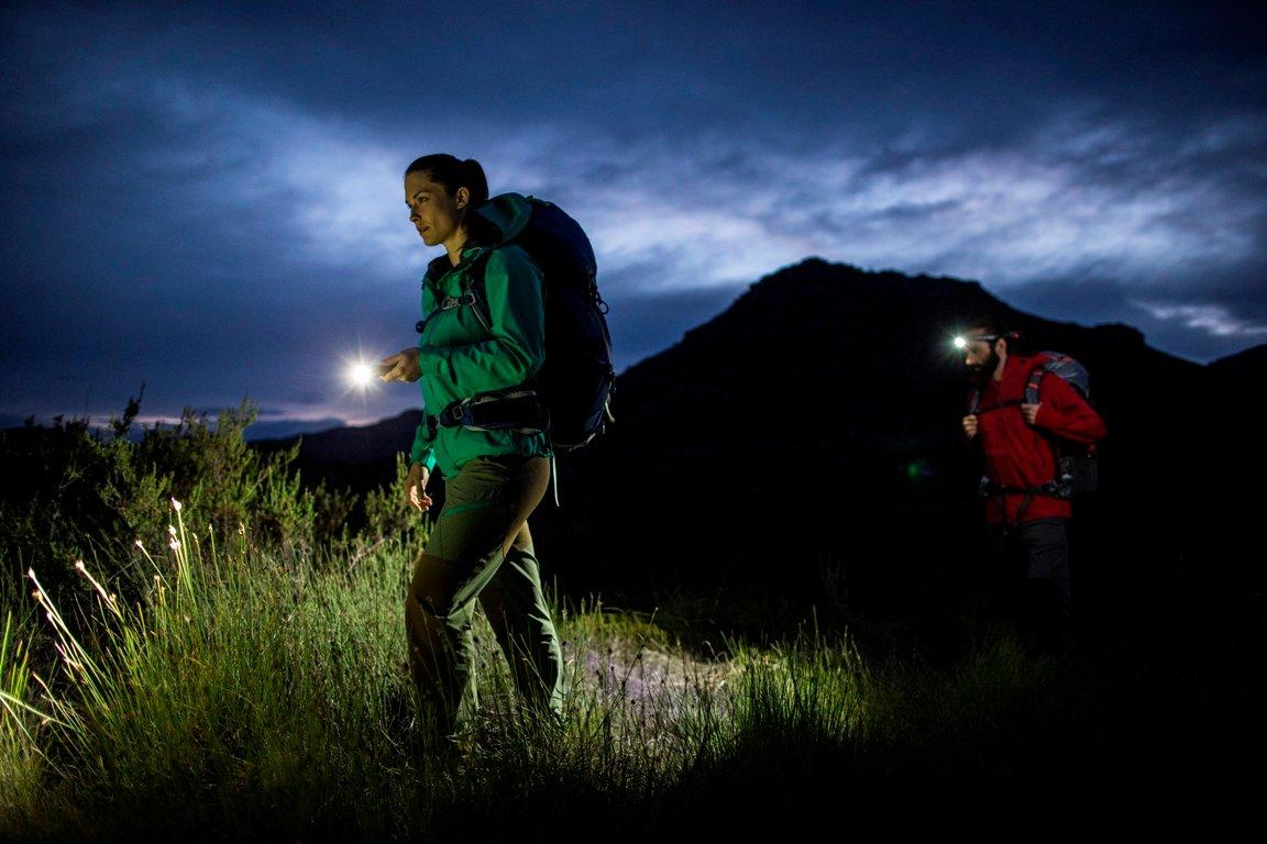 Two people on an evening hike: one is holding a torch and the other has a head torch. In the background is a mountain.