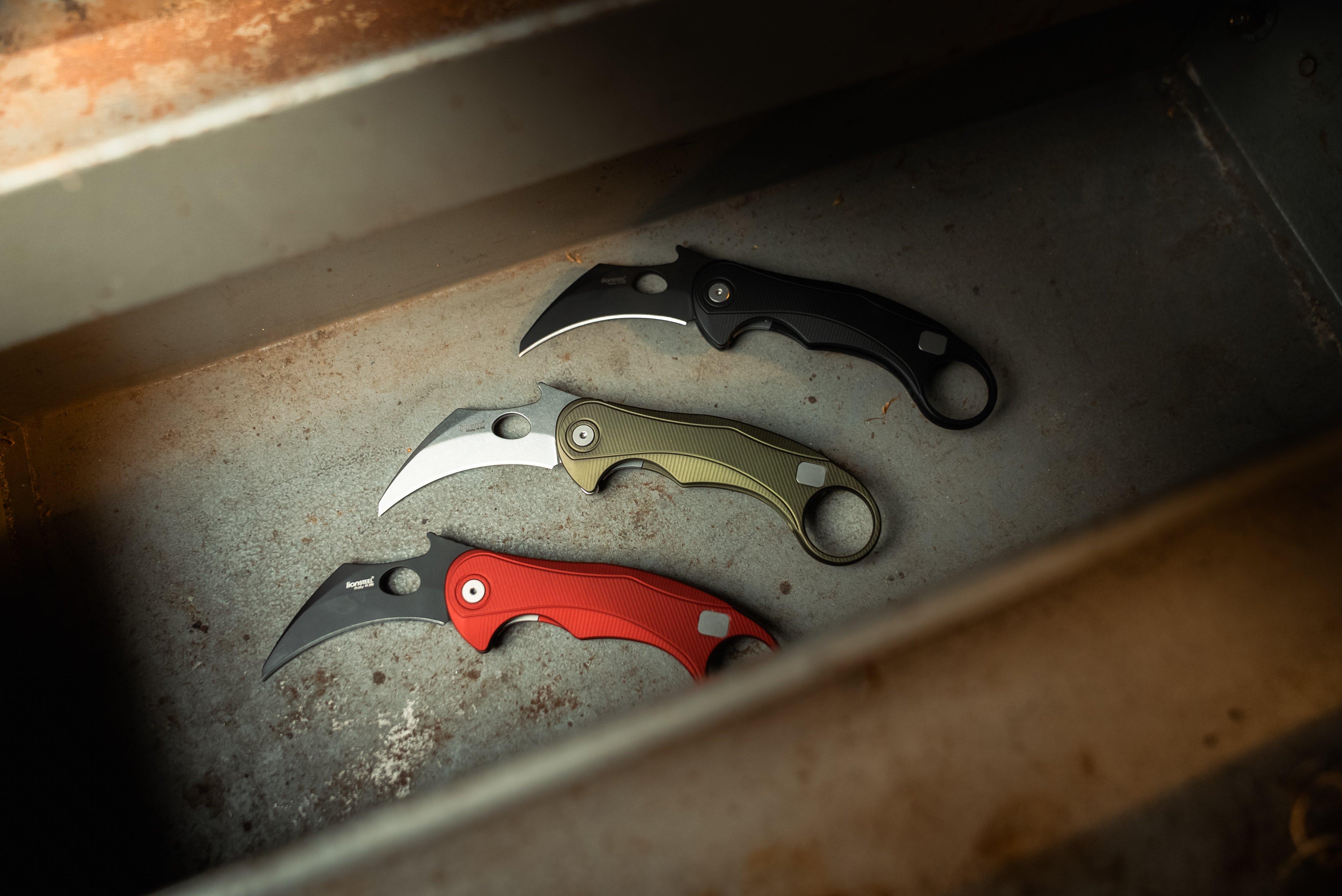 Three Lionsteel L.E.One Mid pocket knives in different colours on a steel surface.