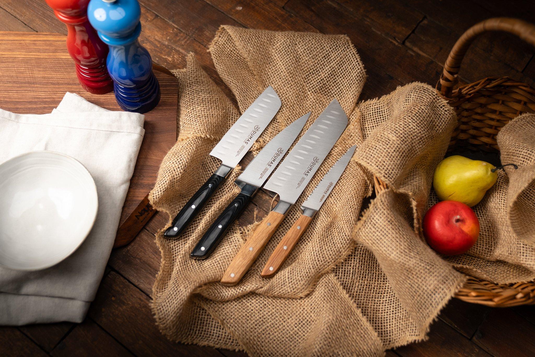 Four 32 Dumas kitchen knives on a wooden table with a jute tablecloth, a blue pepper mill, and a wooden cutting board.