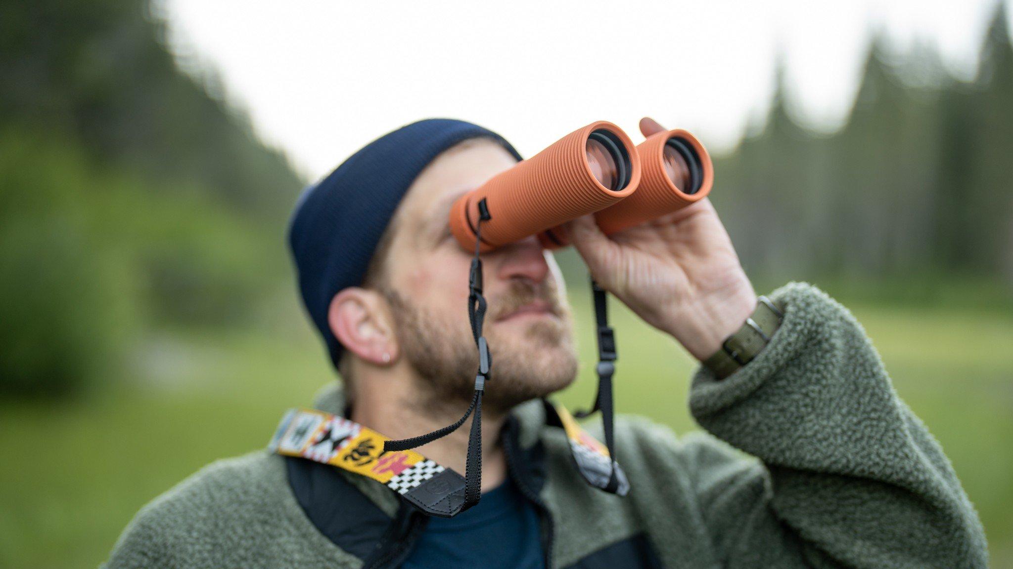 Man in een groen fleecejack die door een paar oranje verrekijkers kijkt in de natuur.