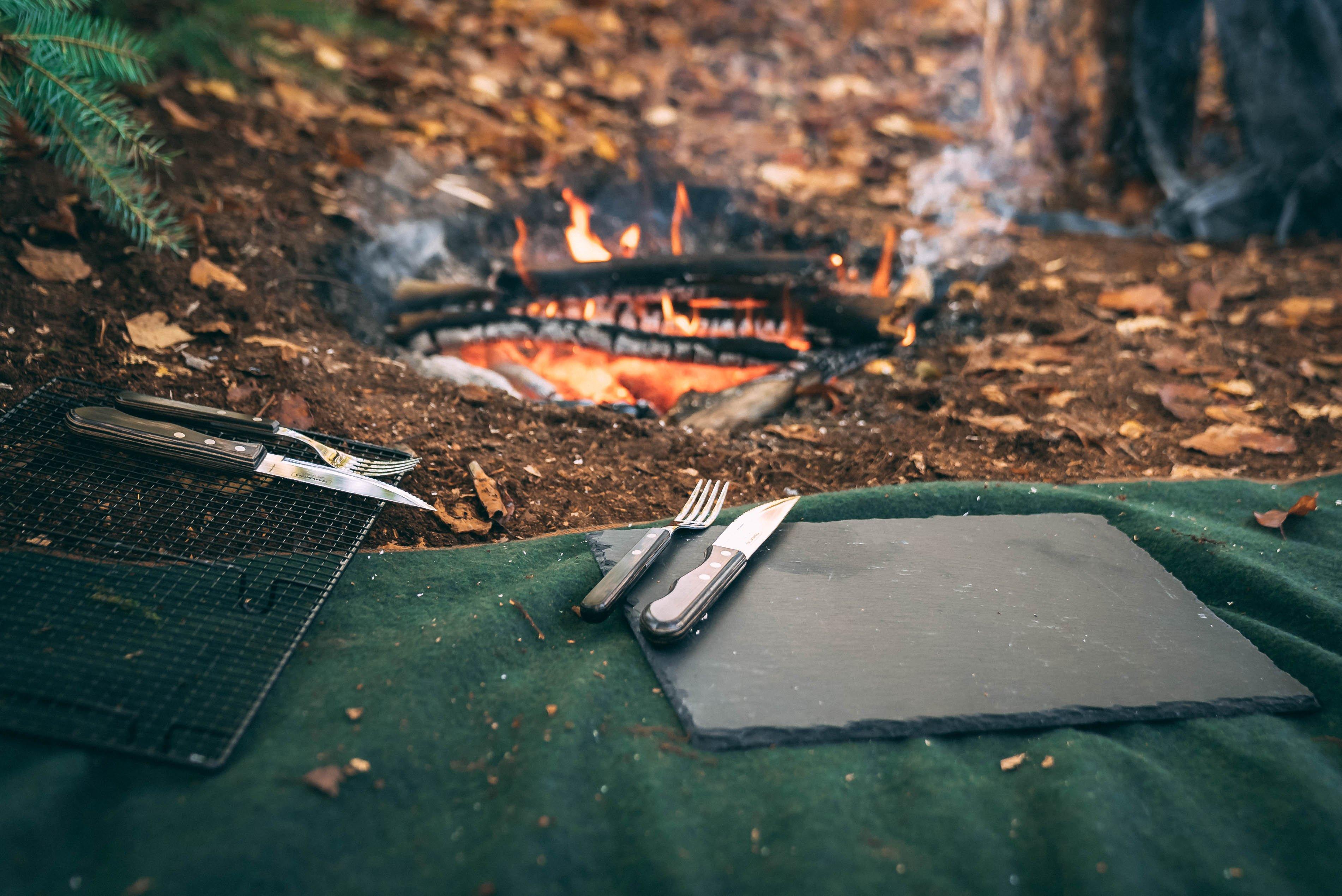 Een set steakmessen en vorken op een leisteen serveerplank, op een bossige ondergrond bij een kampvuur