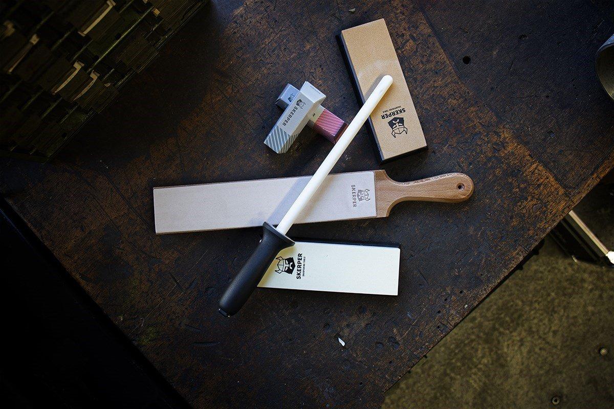 Two Skerper sharpening stones, a strop, stropping compound and a sharpening steel lie on a dark wooden board.