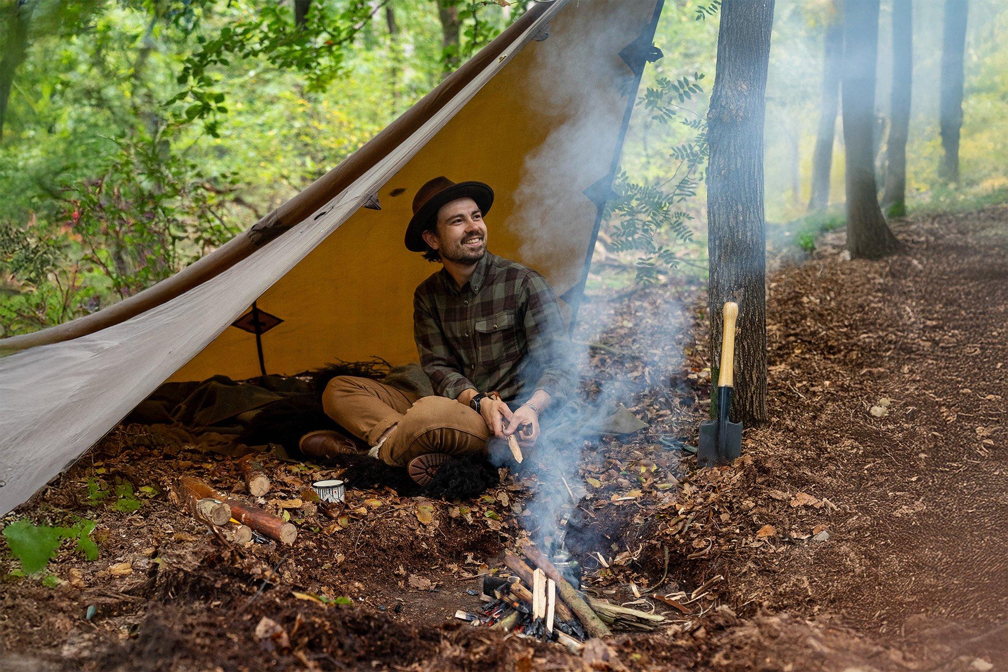 Man zit ontspannen onder tarp in het bos bij een kampvuurtje, omgeven door herfstbladeren en bushcraftuitrusting