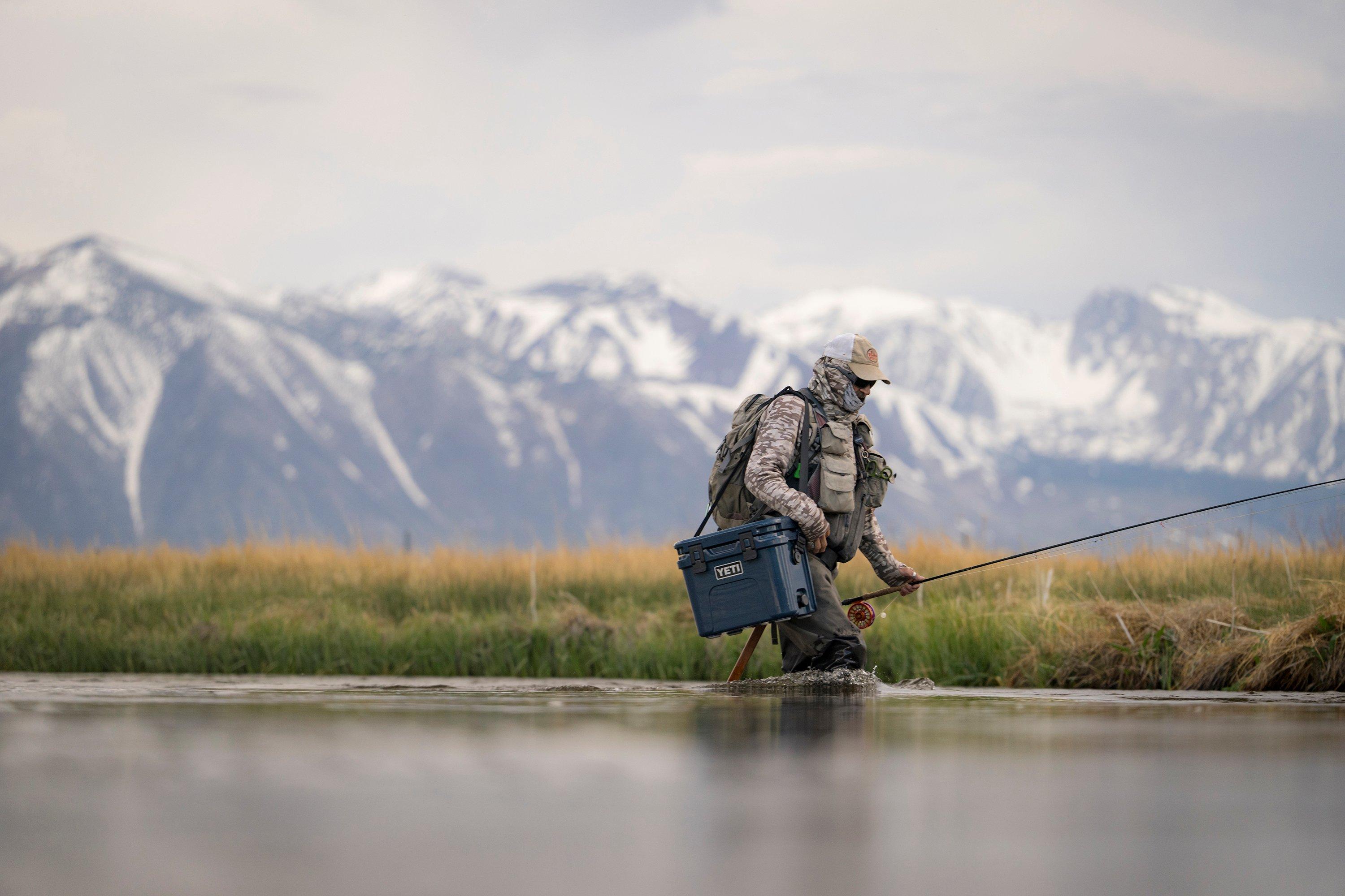 A fly fisherman with a mountain range in the background making his way through a river, carrying a Yeti cooler slung over his shoulder.