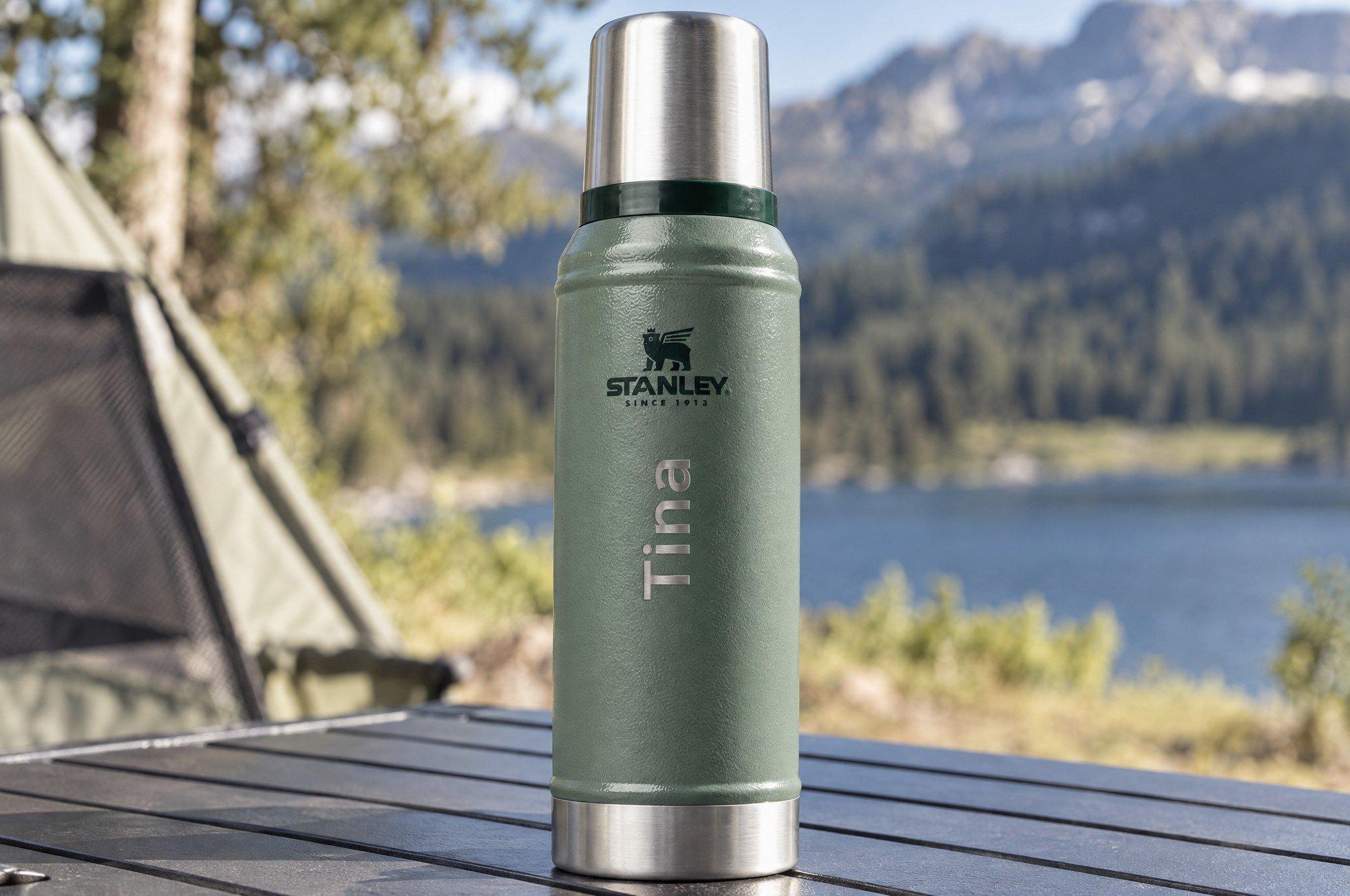 Groene Stanley-thermosfles met de gegraveerde naam “Tina”, op een tafel bij een meer