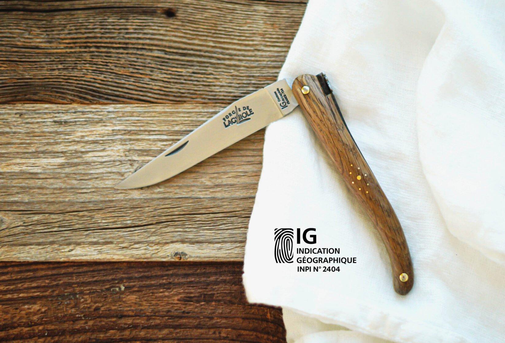 Forge de Laguiole pocket knife on a wooden surface and white cloth, with the Indication Géographique visible on the blade and added as a graphic element in the image.
