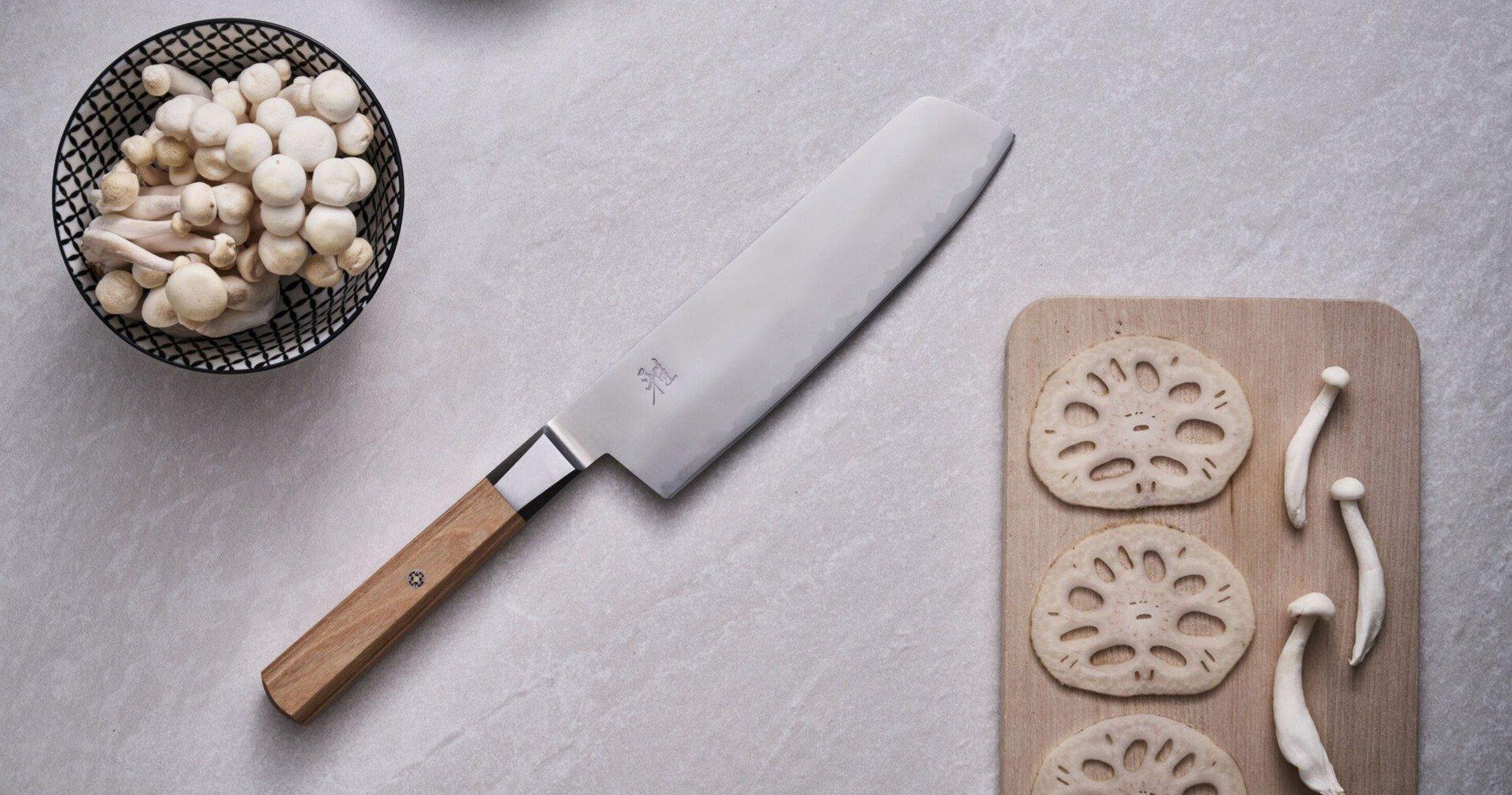 Un nakiri Miyabi by Zwilling KOYA sur une surface grise avec une planche à découper et un bol de champignons blancs.