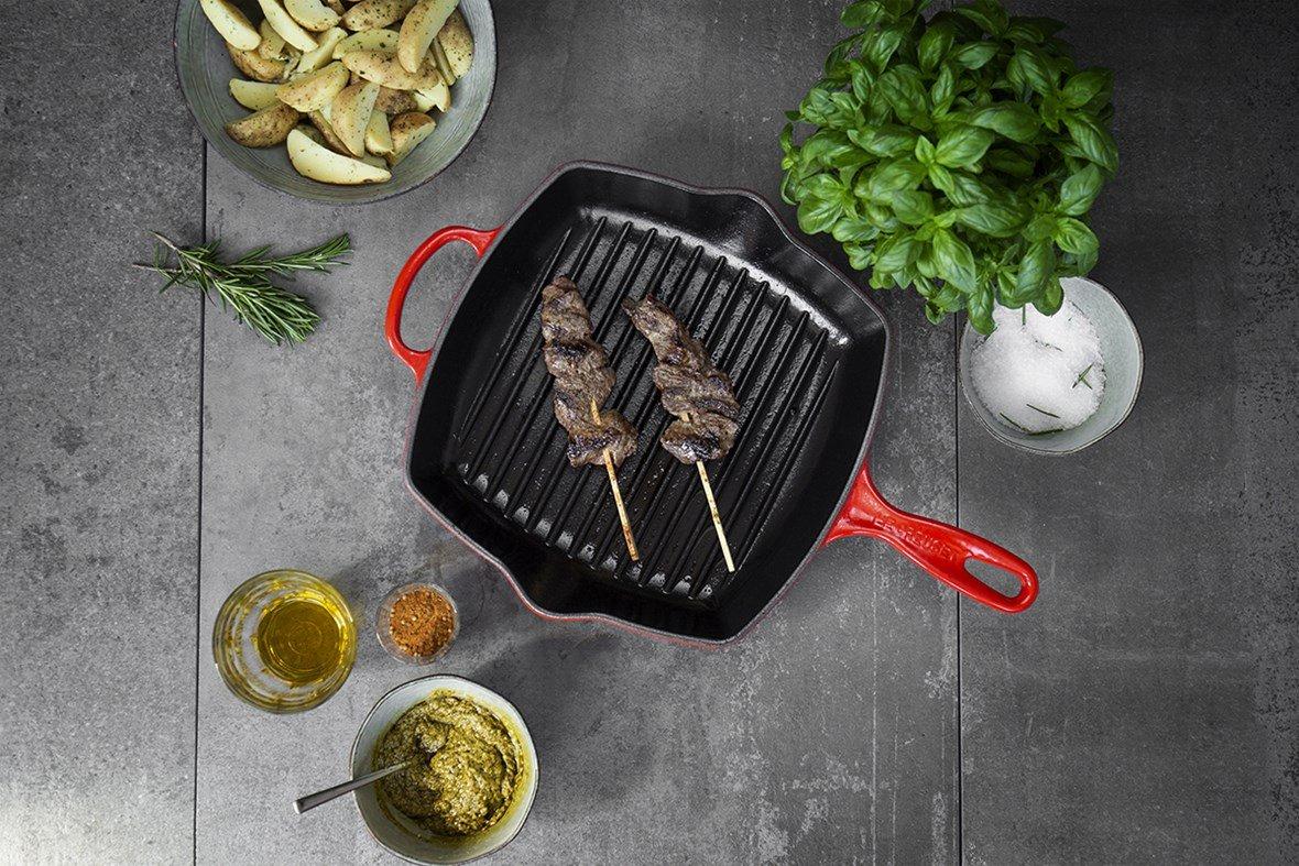 Grelha de ferro fundido vermelha com dois espetos de madeira com carne bovina sobre fundo cinzento